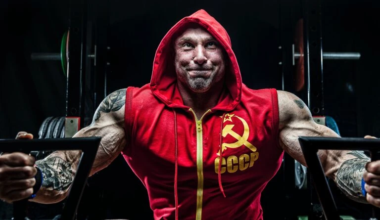 A man in a red hoodie is posing with a barbell.