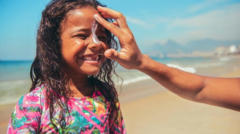 A little girl is being given sunscreen by a man on the beach.